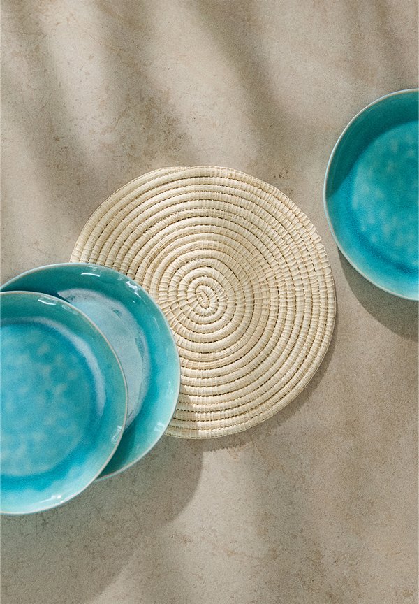 Turquoise ceramic plates piled beside a round, textured woven placemat in a natural colour on a light surface.