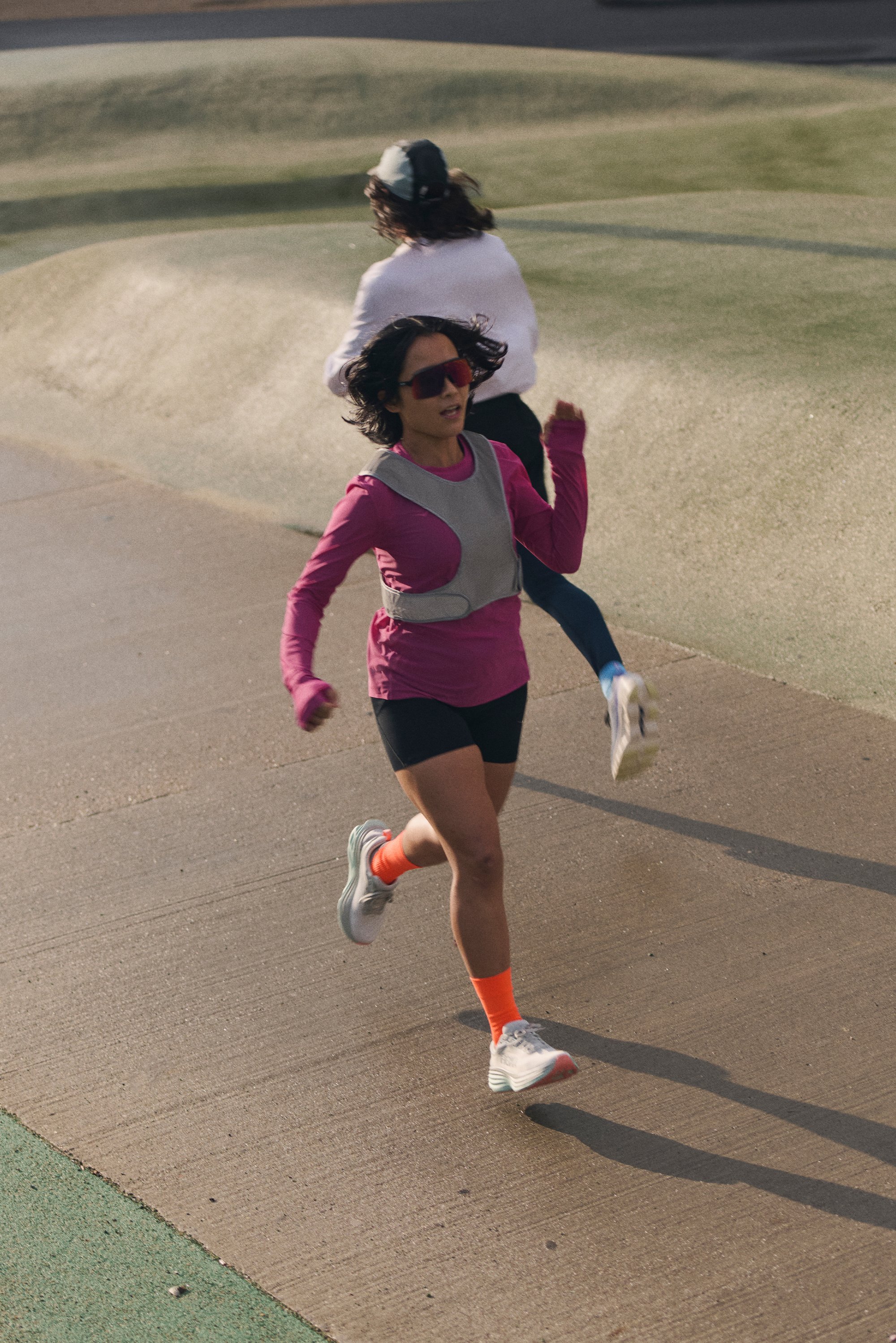Two women run on a path; one wears a pink long-sleeve shirt and sunglasses, while the other is in a white top and a cap, slightly behind.