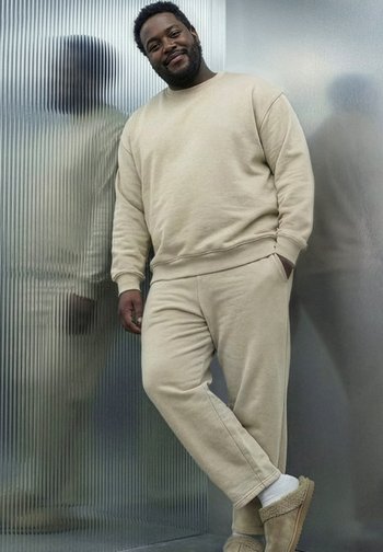 Beige genser- og buksekomplett i mykt, fleece-materiale. Buksene har sidelommer. Modellen poserer foran en teksturert, reflekterende bakgrunn.