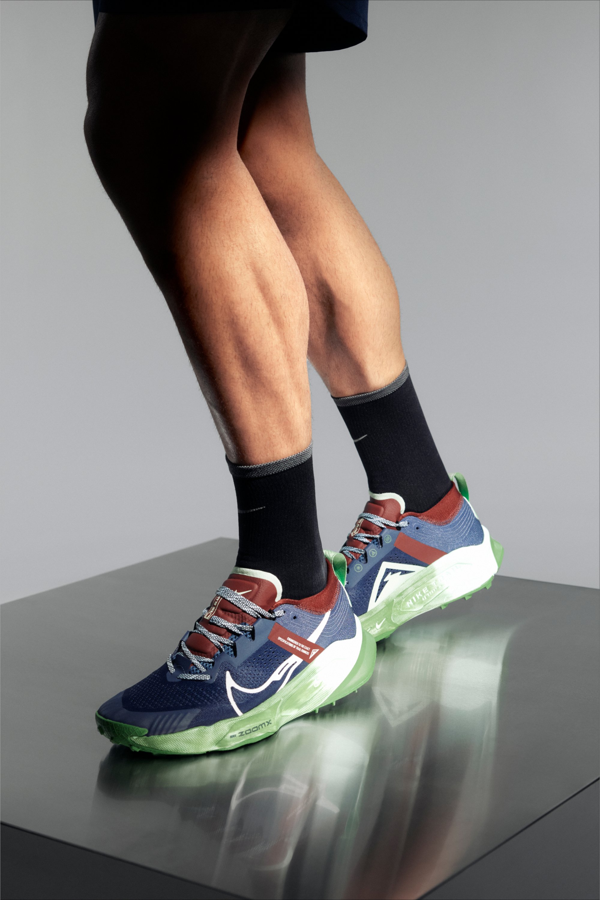 Un primer plano de las piernas de una persona que lleva zapatillas deportivas de colores y calcetines negros, de pie sobre una superficie reflectante con un fondo neutro.