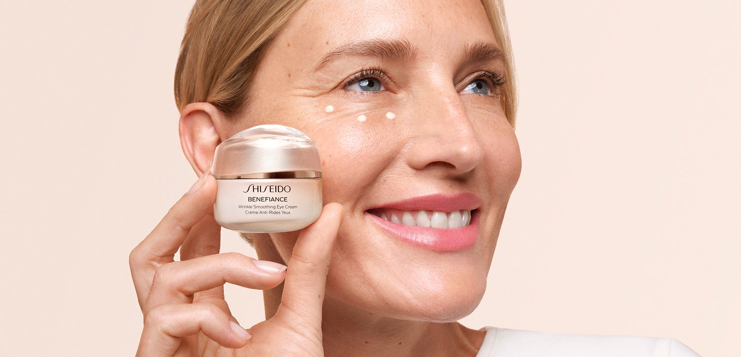 Kobieta uśmiecha się, trzymając krem pod oczy Shiseido Benefiance Wrinkle Smoothing blisko twarzy, z kroplami kremu pod oczami.