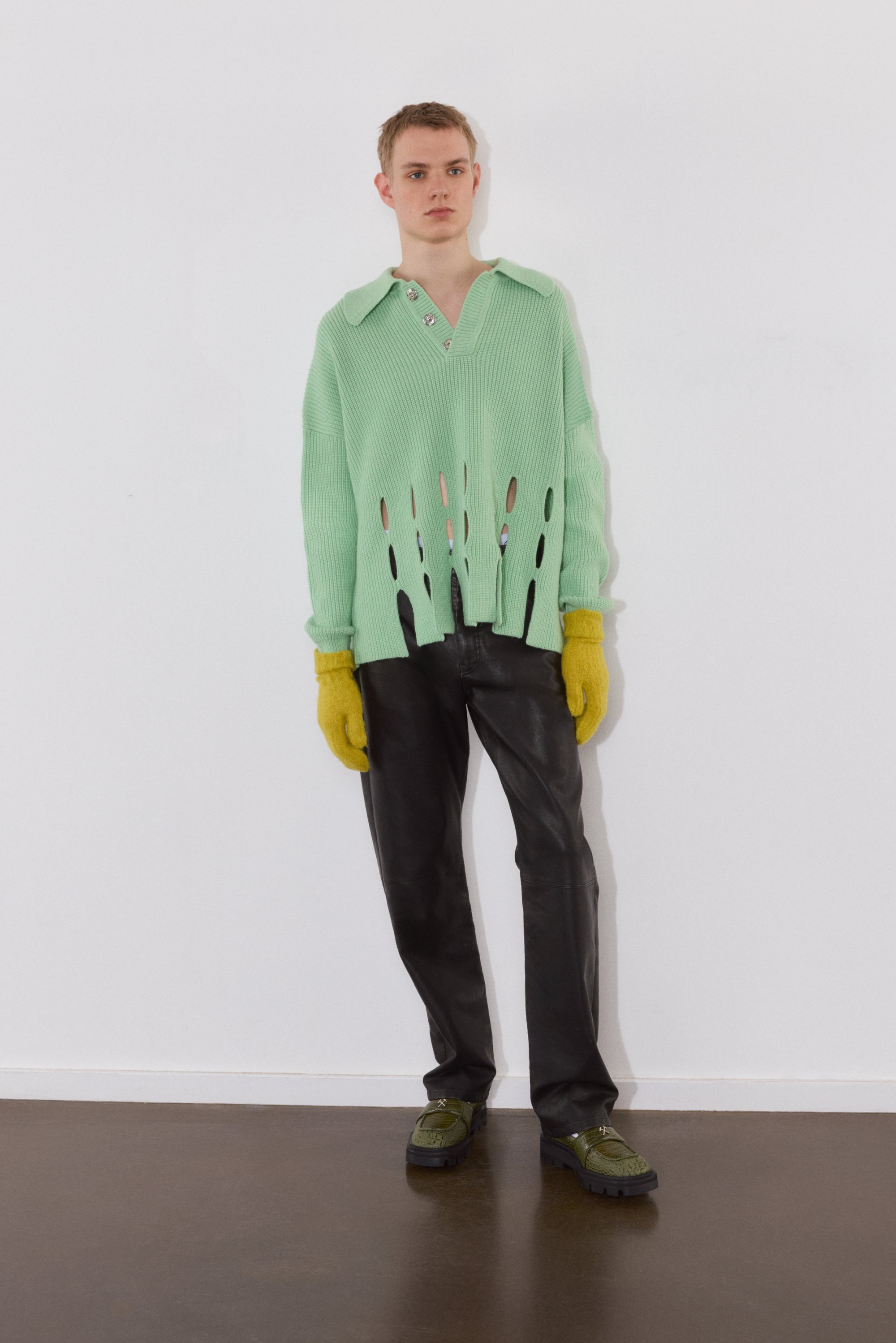Una joven modelo se sostiene contra una pared blanca, vistiendo un suéter oversized verde menta con cortes, pantalones negros, guantes amarillos y zapatos verdes.