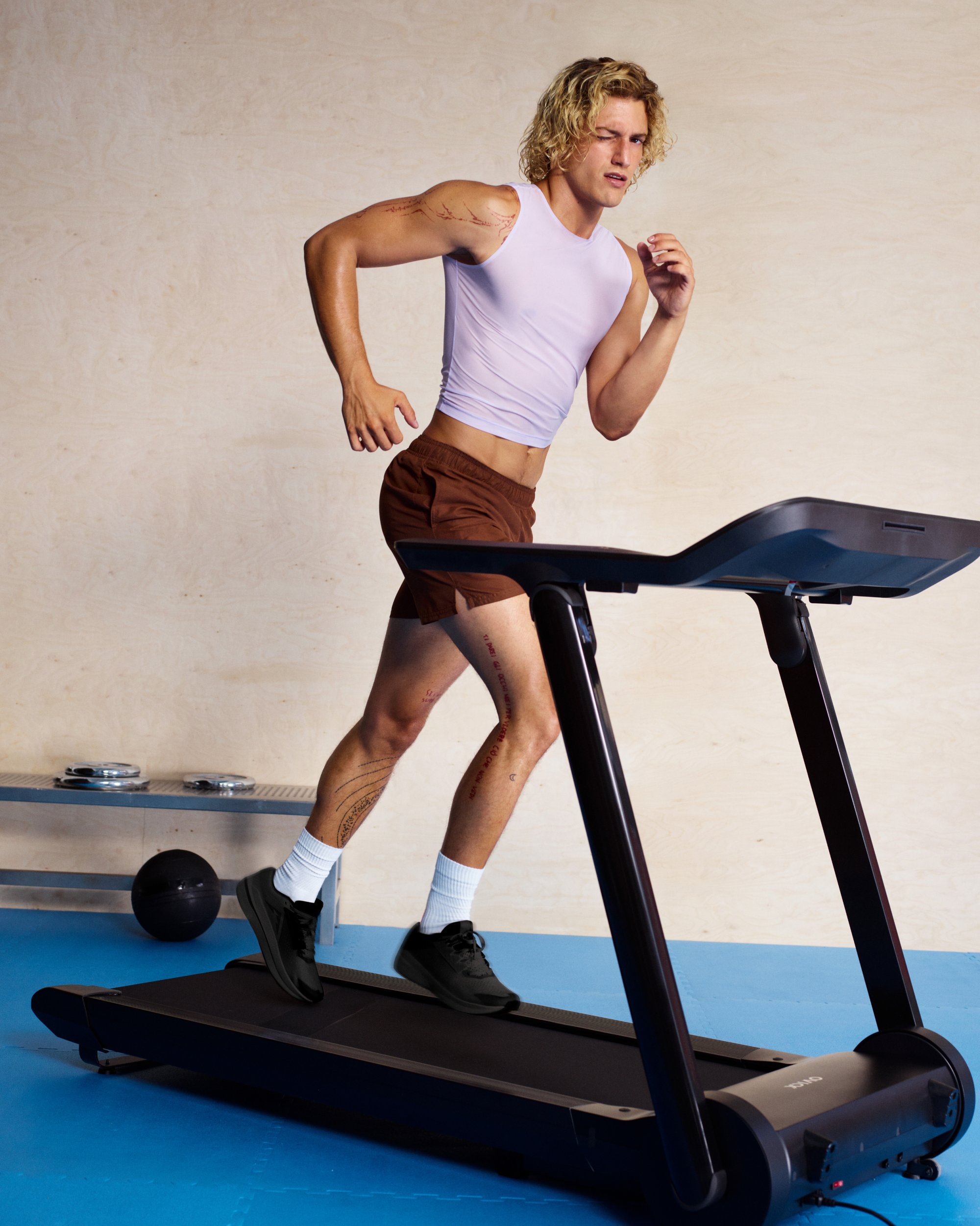 A young man with curly blonde hair runs on a treadmill, wearing a light purple tank top and brown shorts, in a bright gym setting.