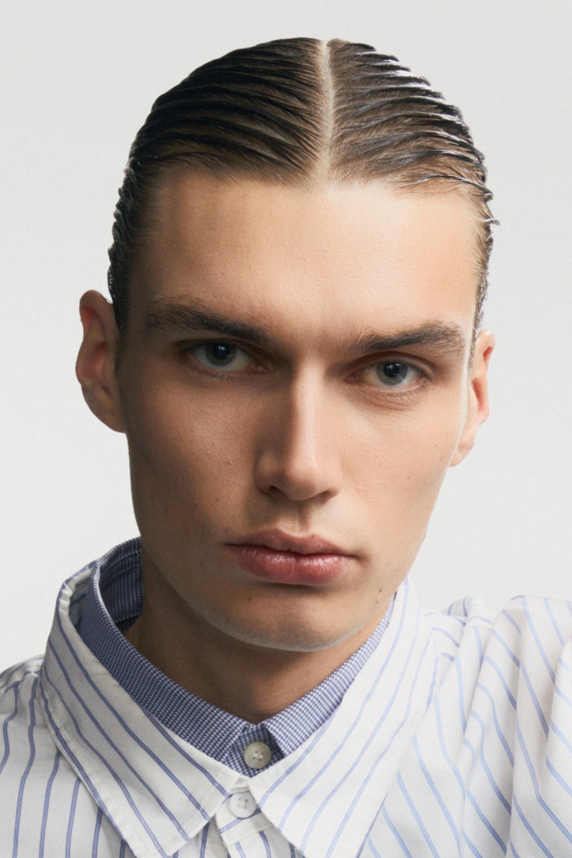 Camisa blanca con rayas verticales azules, cuello con cuadros azules en contraste. Piel suave, cabello peinado hacia atrás, expresión neutral.