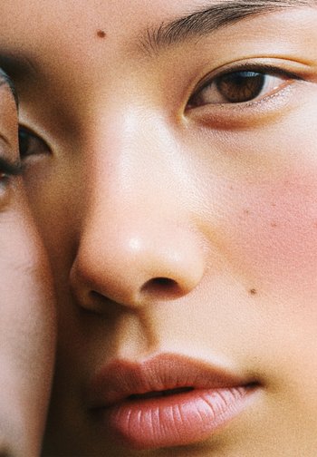 Close-up of a face with smooth, light skin; brown eyes; full lips; subtle blush; a small mole on the cheek; soft highlights on the skin.