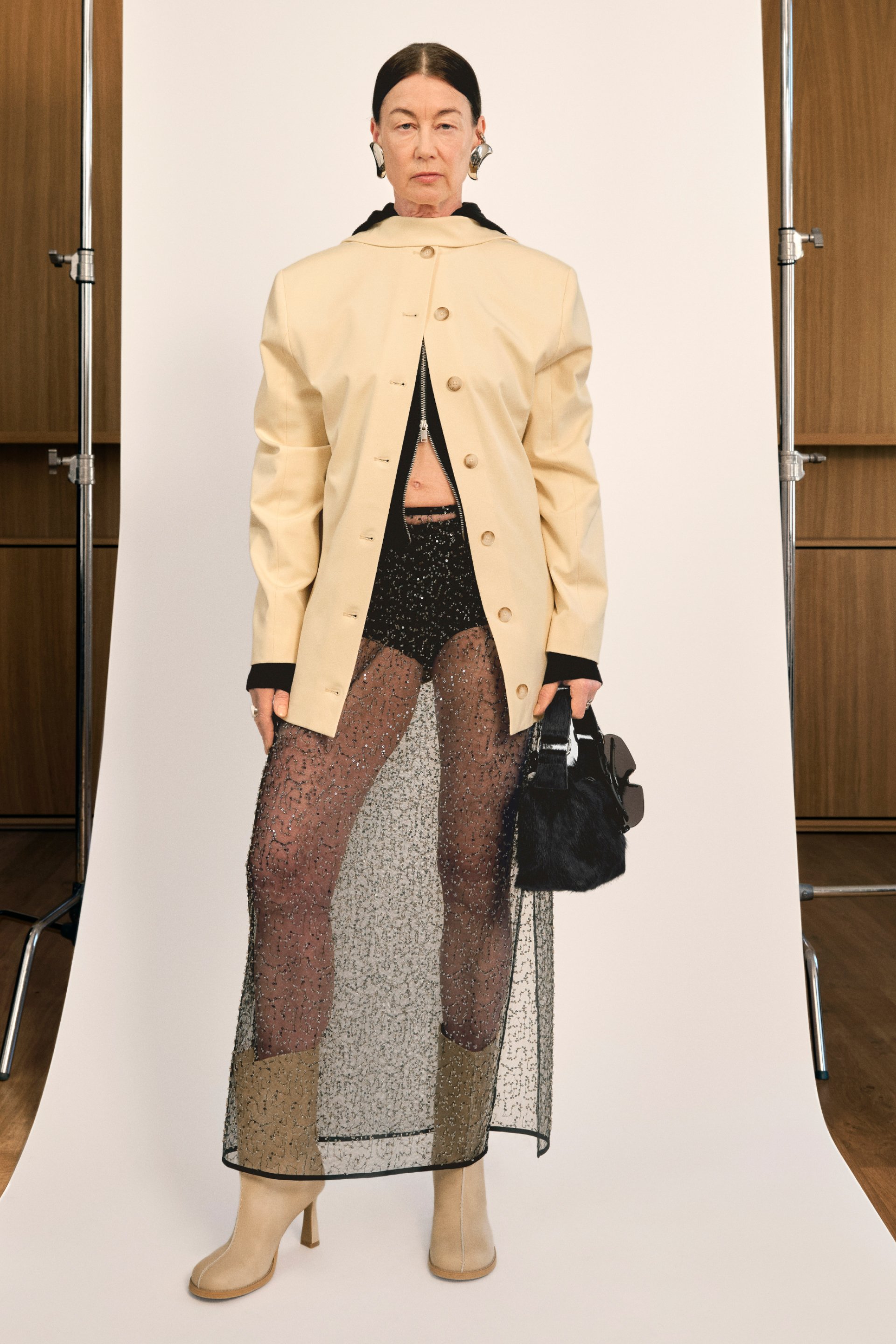 Una modelo se muestra con confianza en un abrigo crema sobre una falda negra transparente, exhibiendo una moda audaz con botas beiges hasta la rodilla y un pequeño bolso negro.