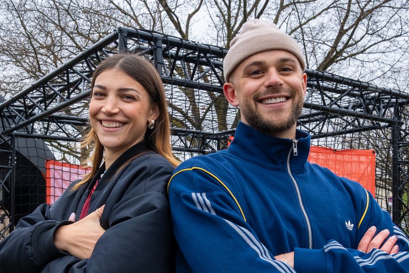 Two smiling adults with arms crossed stand back-to-back outdoors, with a metal structure and leafless trees in the background.