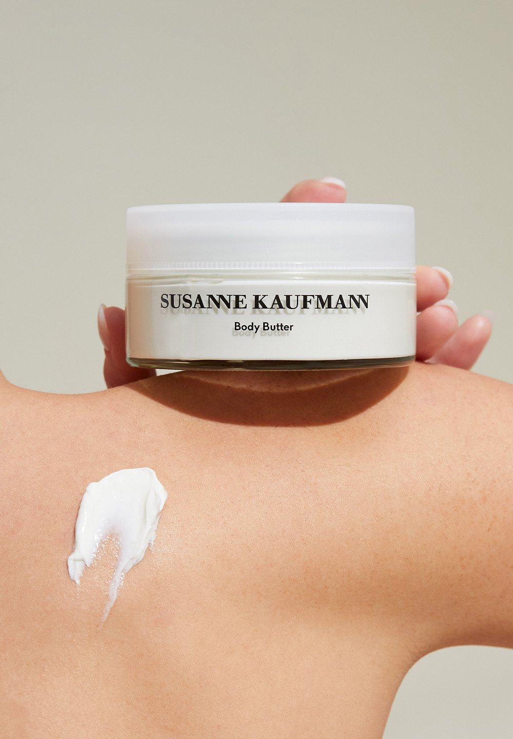 Vit burk med body butter från Susanne Kaufmann, med ett genomskinligt lock och etikett, placerad på hud. Krämig konsistens provad på axeln.