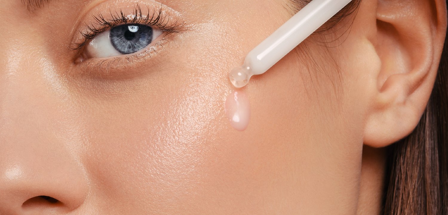 Close-up of a woman's face, highlighting her blue eye and glowing skin, as a drop of serum falls from a dropper onto her cheek.