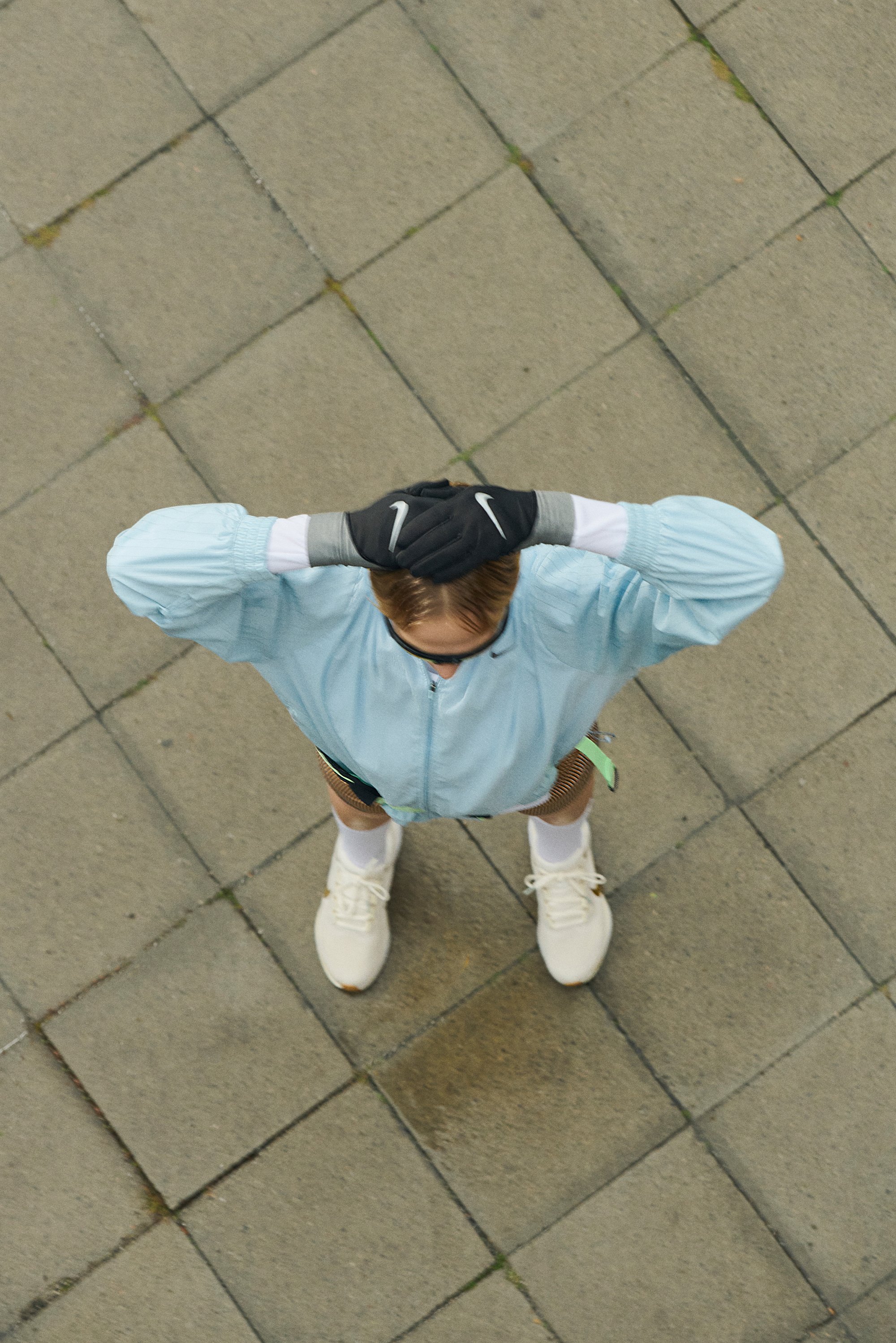 A person wearing a light blue jacket, black gloves, and shorts stands on a stone pavement, hands on head, looking up.