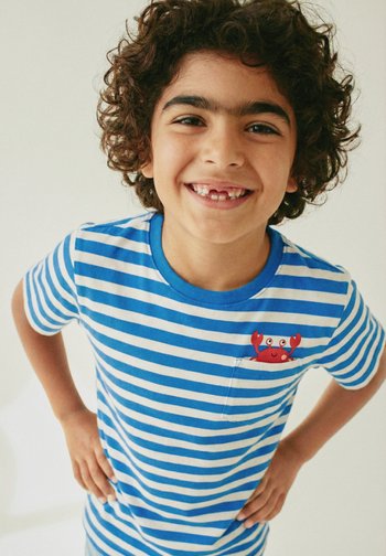 Garçon souriant aux cheveux bouclés avec des dents de devant manquantes, portant une chemise rayée bleu et blanc avec un motif de crabe rouge sur la poche poitrine.