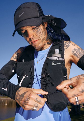 Tatoveret person iført sort Nike-kasket og hydreringsvest justerer vesternes snore udendørs under klar blå himmel.