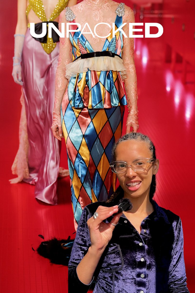 Kleurrijke, gestreepte jurk met diamanten ontwerp, aangevuld met kantaccenten. Model in een zacht marineblauw fluwelen top houdt een kleine borstel tegen een rode achtergrond.