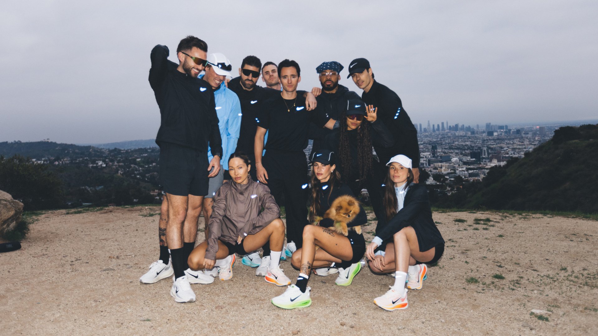 Grupo de individuos diversos en ropa deportiva posando al aire libre con un horizonte urbano de fondo, un perro pequeño en los brazos de una persona.