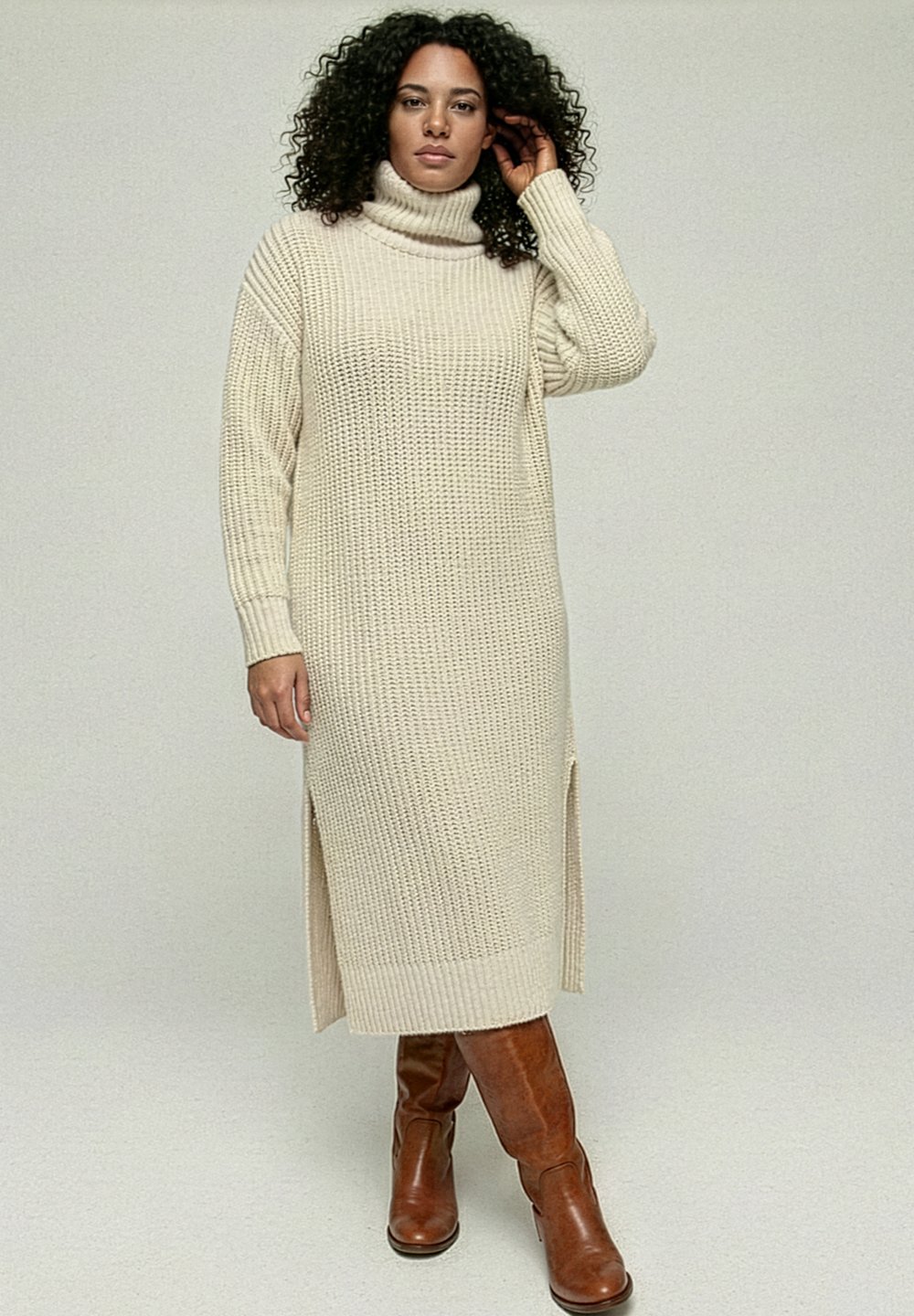 Roomkleurige gebreide coljurk met geribbelde structuur, zijopeningen en lange mouwen, gecombineerd met bruine kniehoge laarzen.