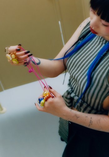 Una persona sostiene anillos grandes y coloridos en una mano y unos auriculares rosa en la otra, mostrando un cabello vibrante y una camisa a rayas.