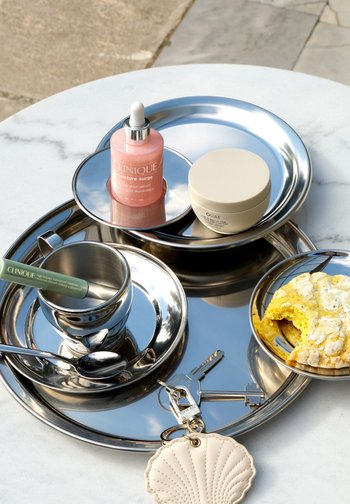 Heijastavat metalliset tarjoiluvadit pitävät Clinique-seerumia ja ripsiväriä, voidepurkin, kupin lusikan kanssa, puoliksi syödyn keltaisen keksin ja avainrenkaan, jossa on kaksi avainta.