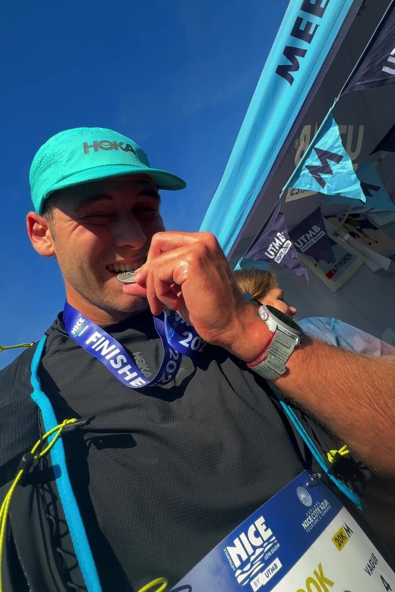 Běžec v černém tričku s tyrkysovými doplňky, s tyrkysovou čepicí, kousá do stříbrné medaile na modré stuze označené "FINISHER 2023."