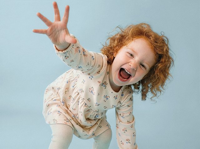Petite fille rousse en robe fleurie tendant la main en riant devant un fond bleu clair uni.