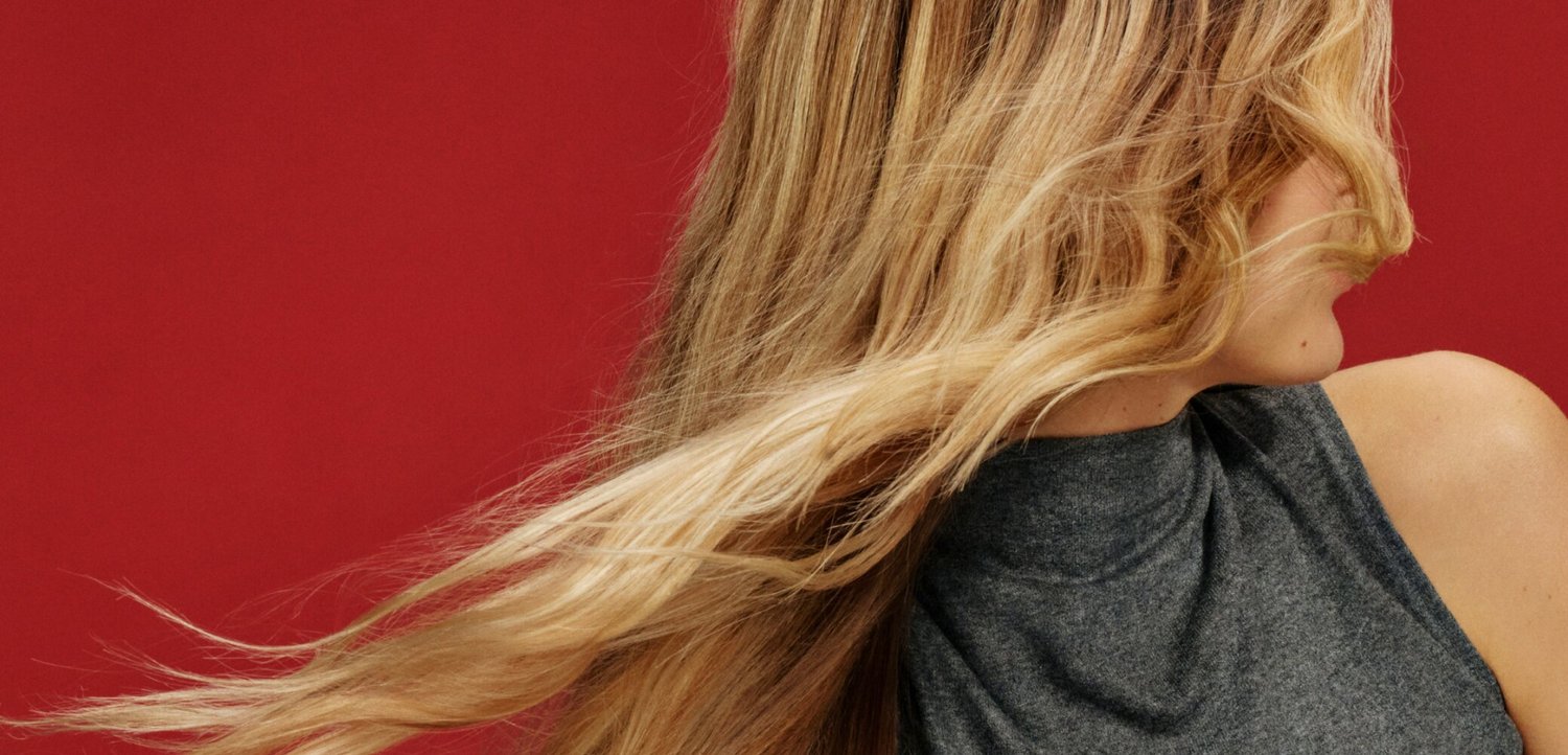 Donna bionda con capelli lunghi girata di spalle, indossa un top grigio scuro senza maniche su uno sfondo rosso uniforme.