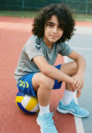Lockiger Junge in Sportkleidung sitzt auf einem Volleyball auf einem Außenplatz, trägt blaue Shorts, graues Shirt, weiße Socken und blaue Laufschuhe.