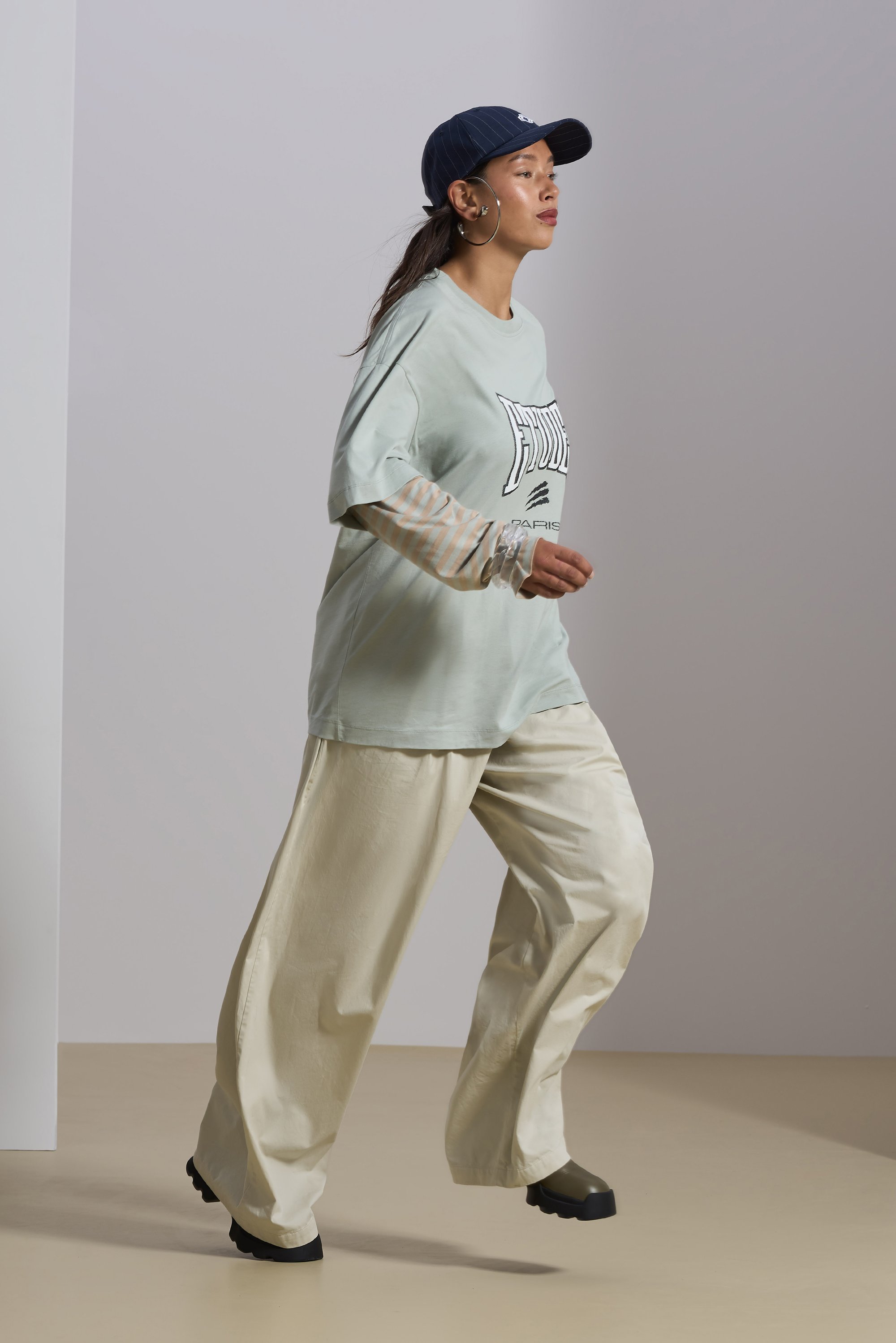 A person walks confidently in a light green oversized shirt and cream wide-leg pants, accessorized with a navy cap and hoop earrings.