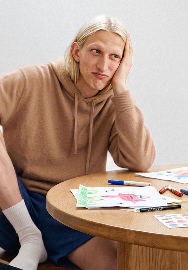 Persona con pelo largo y rubio que lleva una sudadera marrón con capucha, sentada en una mesa con dibujos y rotuladores de colores, apoyando la cabeza en una mano.