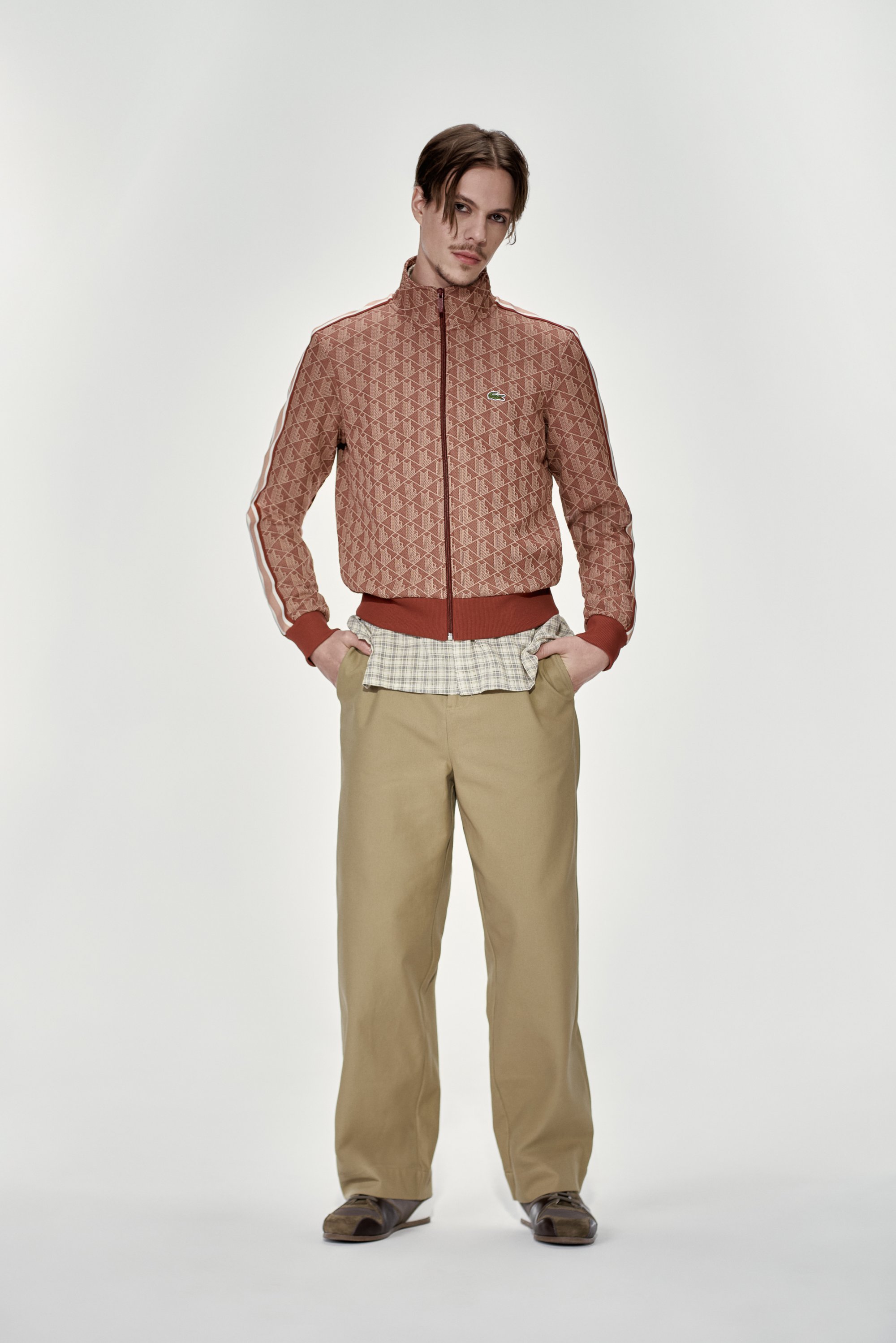 El modelo se muestra con confianza en una chaqueta roja con estampado, una camisa de cuadros y pantalones beige, con calzado marrón, frente a un fondo blanco liso.