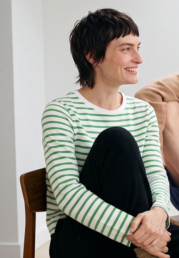 Mujer con el pelo corto y oscuro, que lleva una camiseta de manga larga blanca con rayas verdes, sentada con la rodilla levantada, sonriendo y mirando hacia la derecha.