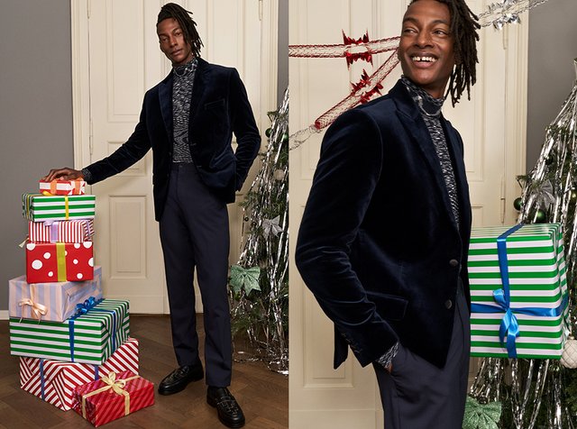 Mann in blauem Samtblazer und gemustertem Rollkragenpullover posiert mit verpackten Weihnachtsgeschenken neben einem geschmückten Baum und einer Tür in einem festlichen Raum.