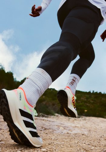 Runner wearing black leggings and white Terrex trainers on a rocky trail with green hills and blue sky in the background.