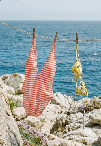 Jednodijelni prugasti kupaći kostim i gornji dio bikinija s uzorkom limuna vješaju se na uže za rublje blizu kamenite obale s plavim morem u pozadini.