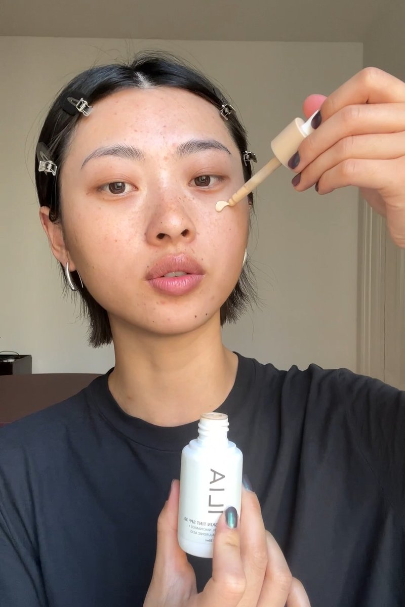 Een persoon met kort haar houdt een druppelflesje vast en brengt vloeistof aan op hun wang. Ze dragen een donker shirt en hebben minimale make-up.