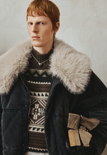 Junge Person mit roten Haaren, die eine dunkle Jacke mit großem Pelzkragen, einen gemusterten Pullover trägt und Brennholz in einer behandschuhten Hand hält.