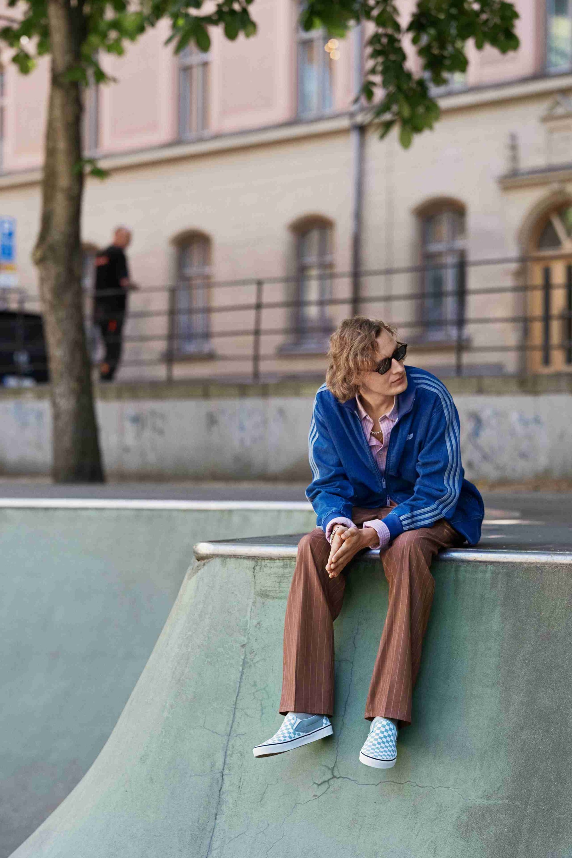 Blue zip-up jacket with three white stripes, light pink shirt, brown striped pants, and blue checkered sneakers, sitting on a skate ramp.