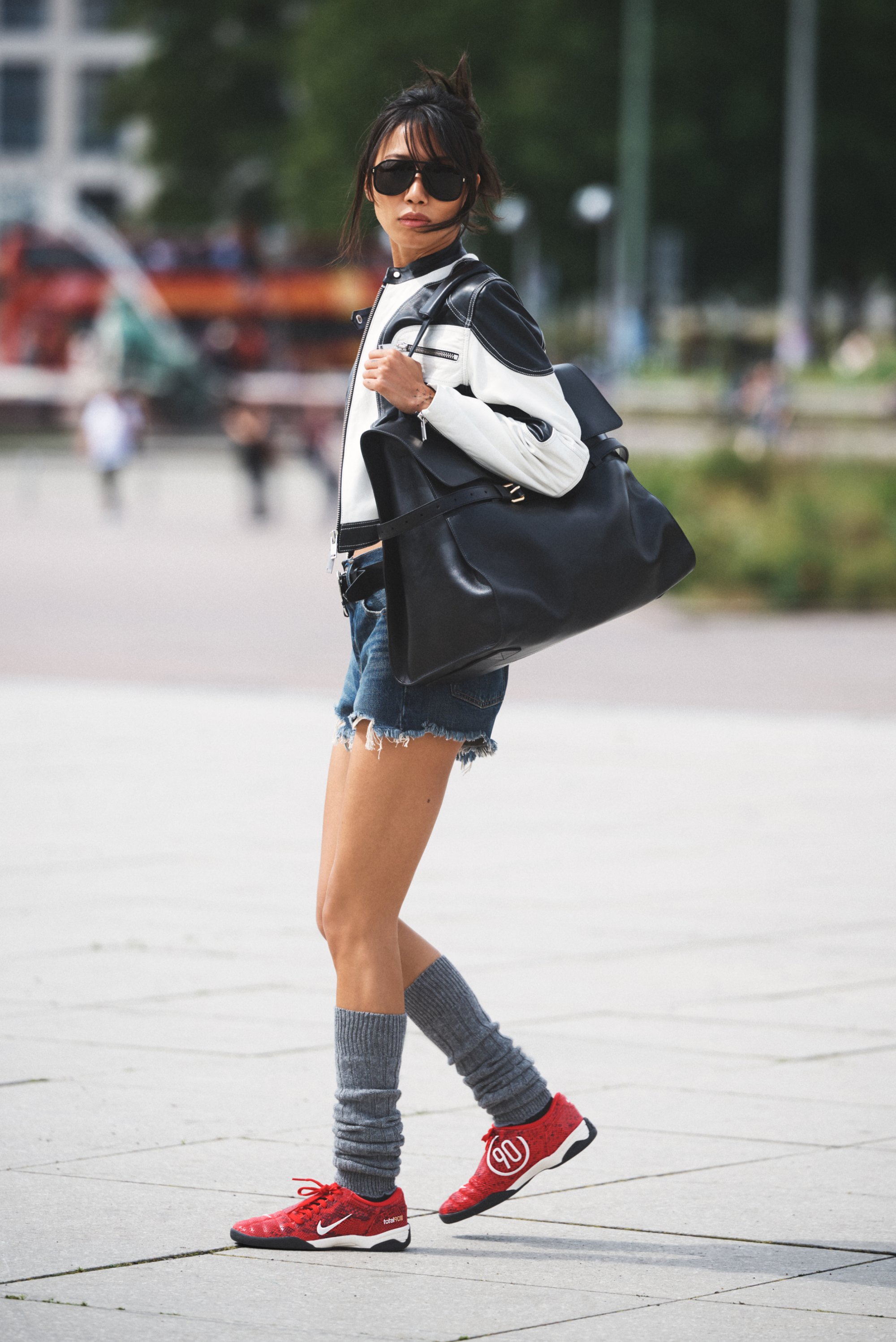 Chaqueta de piel blanco y negro, pantalones cortos de mezclilla, calentadores de piernas grises y zapatillas deportivas rojas. Gran bolso negro, gafas de sol, con un fondo urbano.