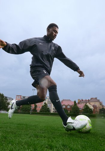 Veste de sport Nike noire et shorts portés par une personne qui frappe un ballon de soccer blanc et vert sur un terrain herbeux sous un ciel nuageux.
