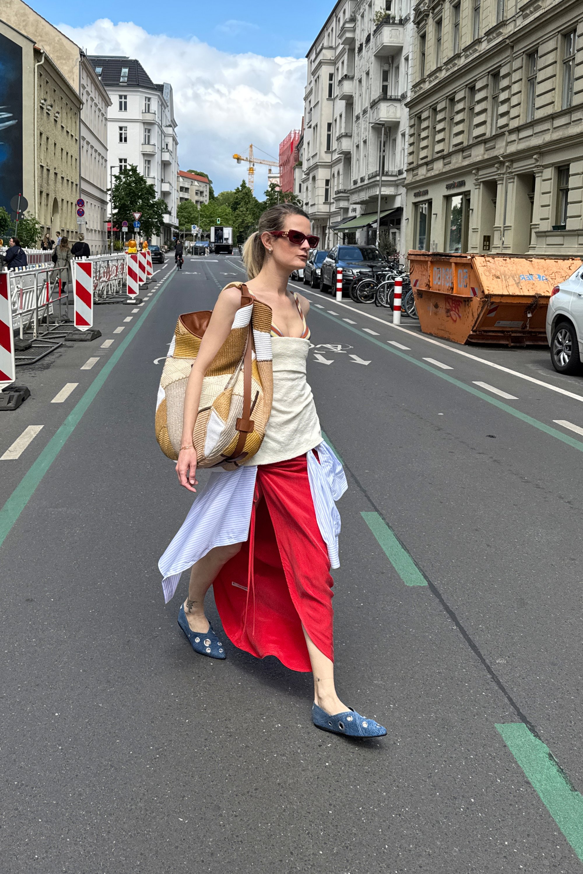 Una donna cammina con sicurezza per la strada, indossando un outfit casual e portando una grande borsa. Sfondo urbano con edifici e un cestino della spazzatura.