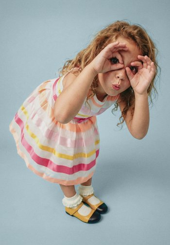 Jeune fille en robe rayée et chaussures jaunes faisant un geste de jumelles avec ses mains, penchée en avant devant un fond bleu uni.