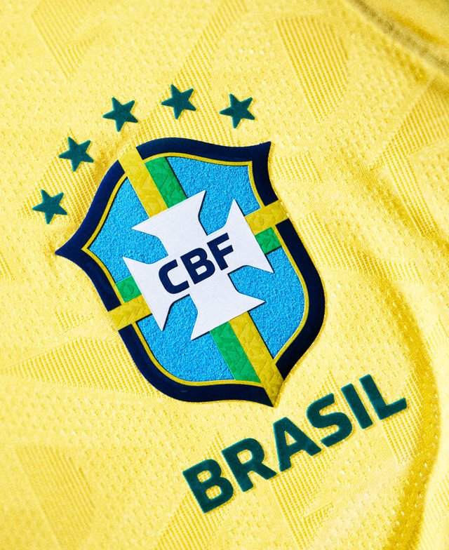 Emblemat reprezentacji narodowej Brazylii w piłce nożnej z pięcioma zielonymi gwiazdami nad nim na żółtym, teksturowanym tle tkaniny.