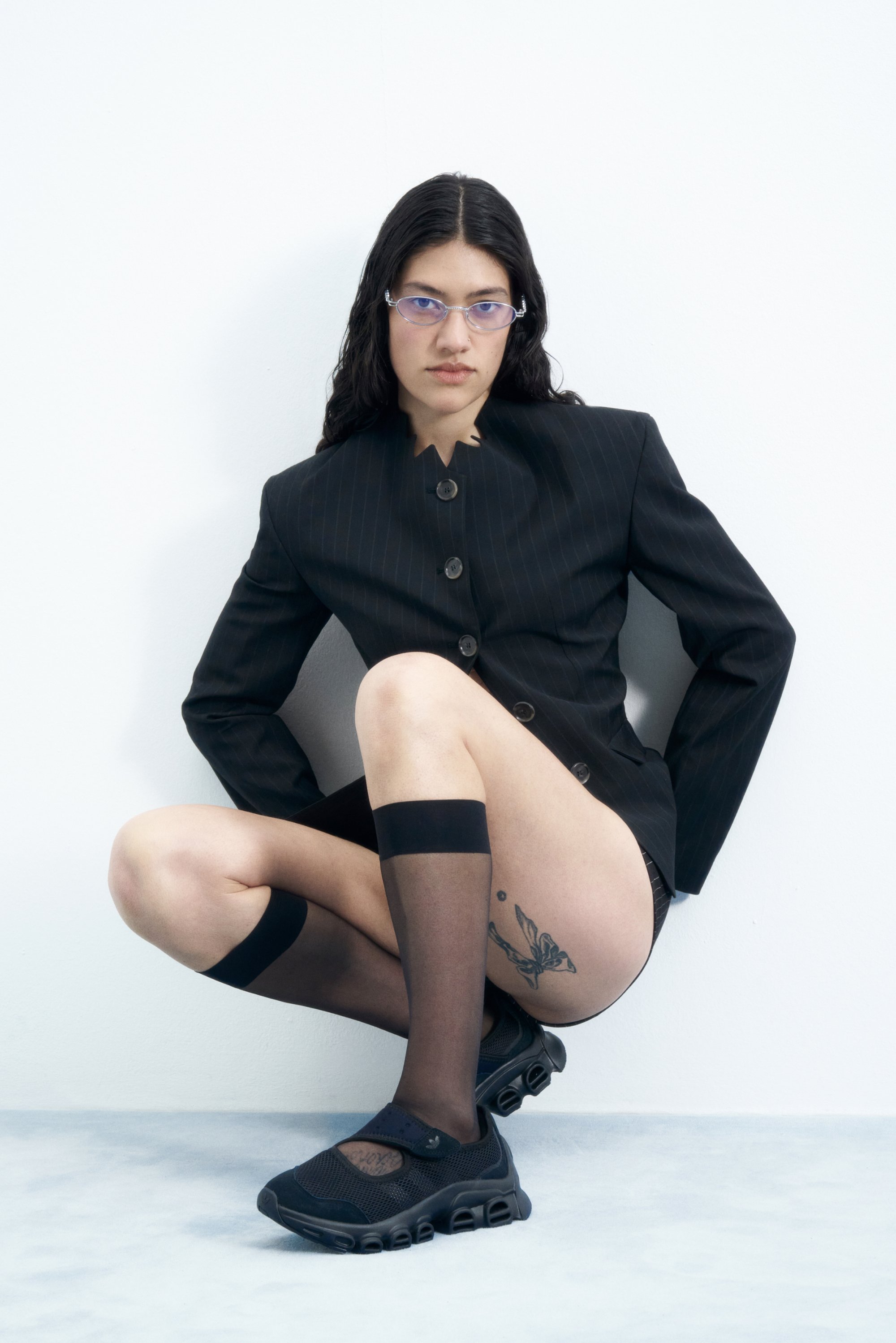 Una persona con cabello largo y oscuro lleva un blazer negro a medida sobre medias hasta el muslo y zapatos deportivos negros, posando frente a una pared blanca.