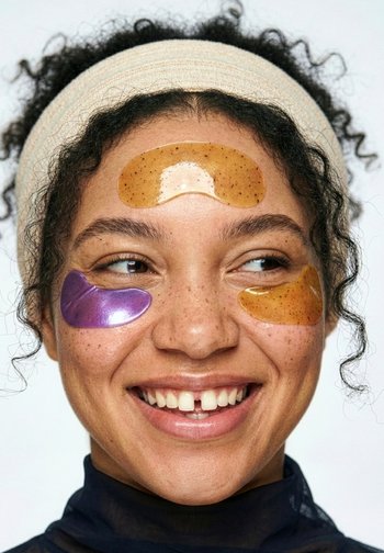 Młoda kobieta z kręconymi włosami, nosząca opaskę oraz kolorowe plastry na twarz pod oczami i na czole, uśmiechająca się i patrząca w bok.