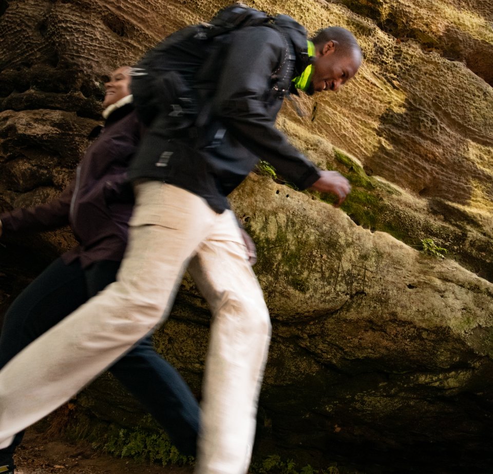 Two people joyfully hiking through a rocky area, with moss-covered cliffs in the background, capturing a sense of adventure and movement.