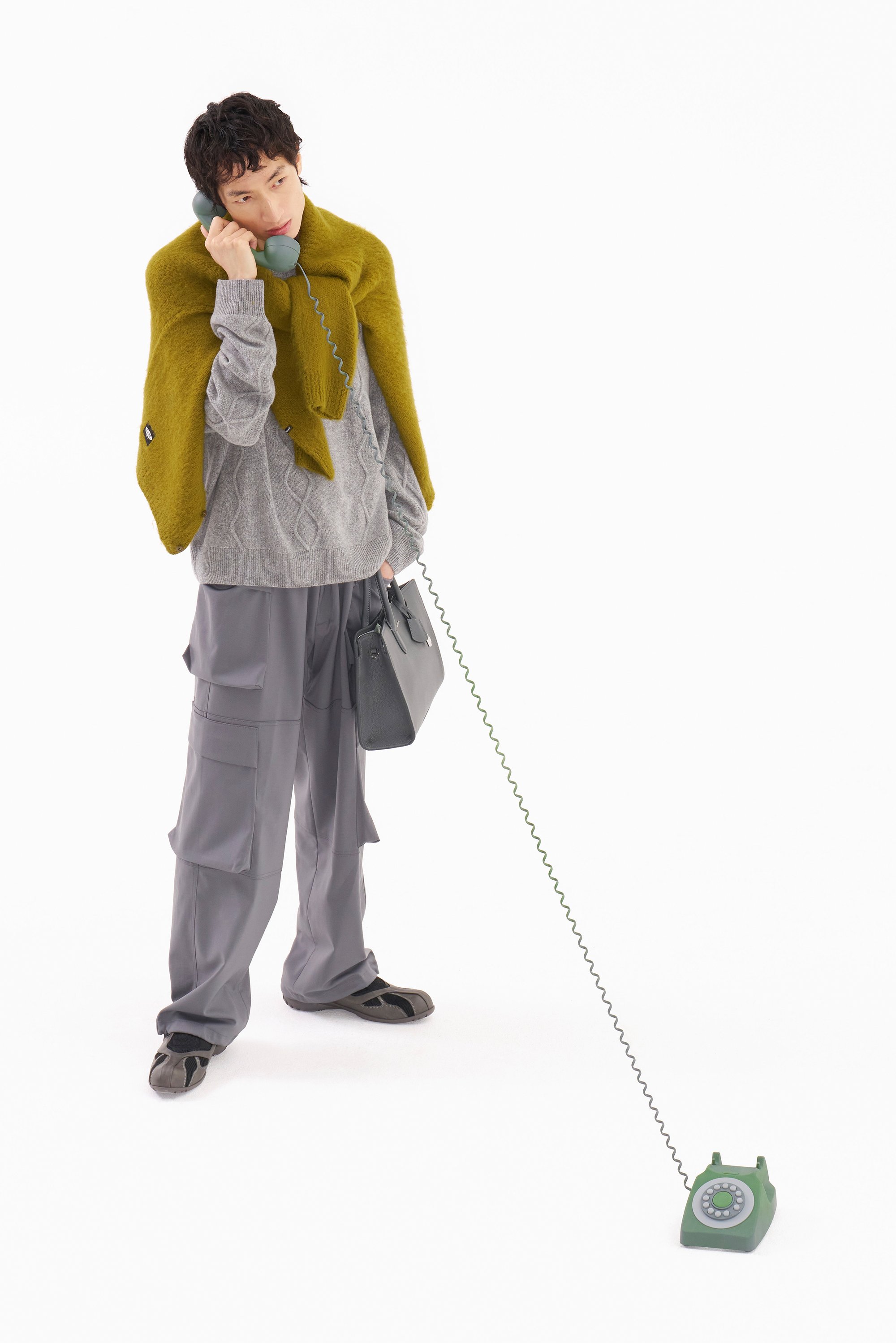 Una persona con un suéter gris y un chal verde habla por un teléfono de disco verde vintage mientras sostiene un bolso gris, con un fondo blanco.