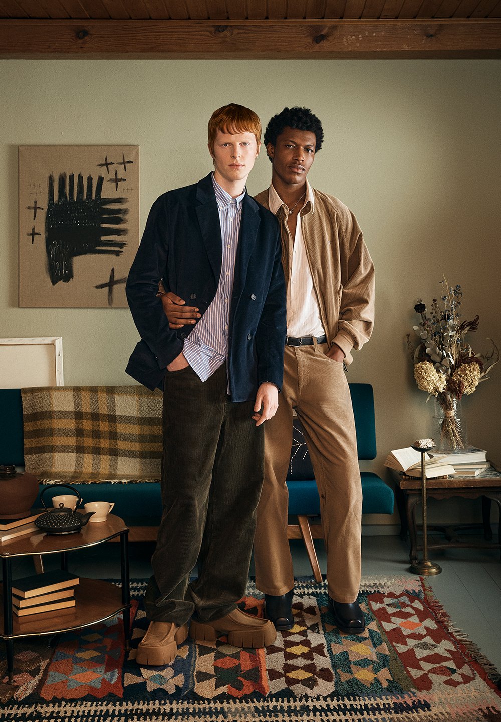 Twee modellen staan samen in een kamer. De ene draagt een marineblauwe corduroy jas, een gestreept overhemd en groene corduroy broek; de ander een beige bomberjack en beige broek.