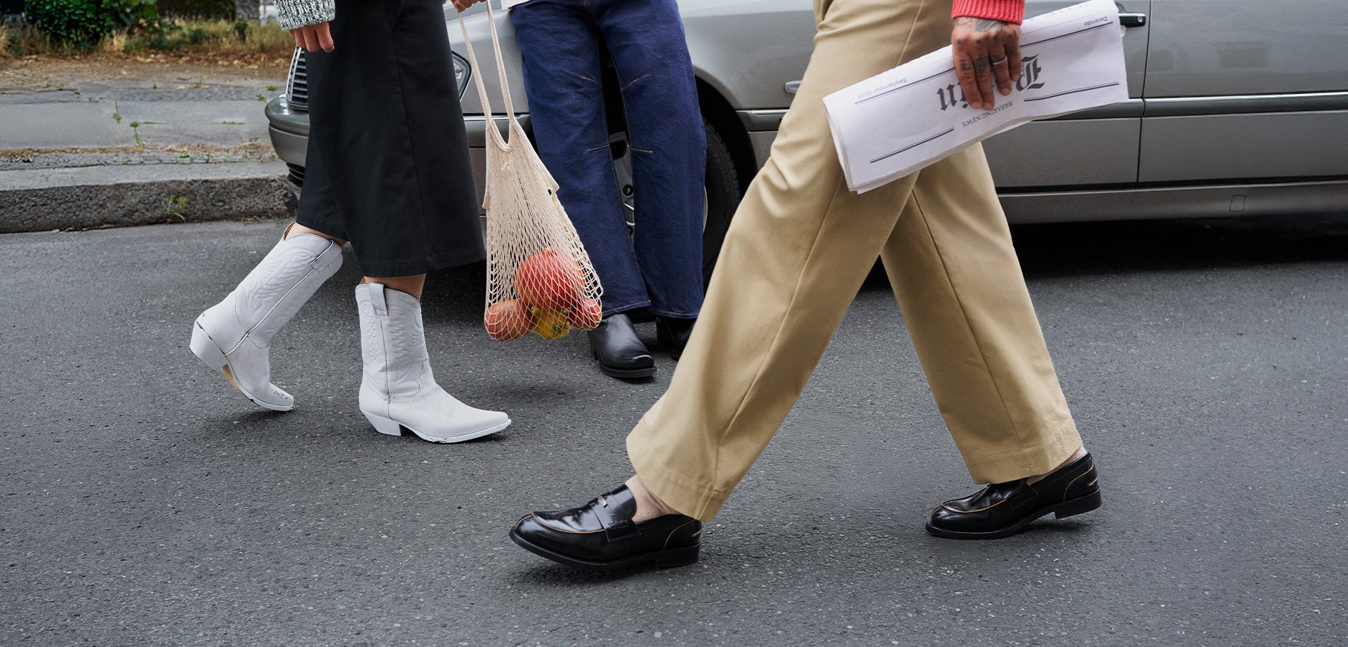 Footwear includes white cowboy boots, black loafers, and tan trousers. A net bag contains apples. The setting is a street next to a parked car.