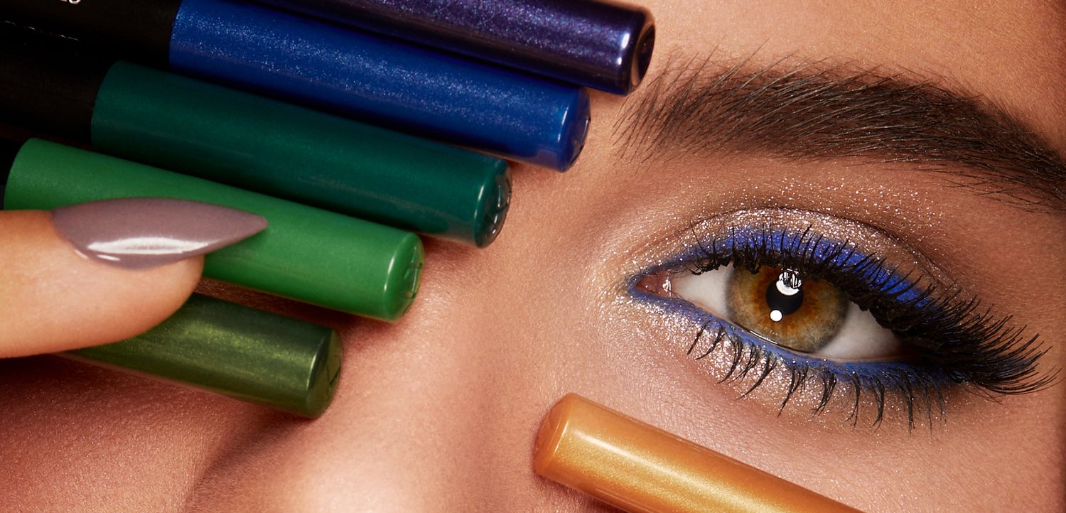 Gros plan d’un œil marron avec un eye-liner bleu vif, de longs cils et une main tenant des crayons de maquillage colorés près de la paupière.
