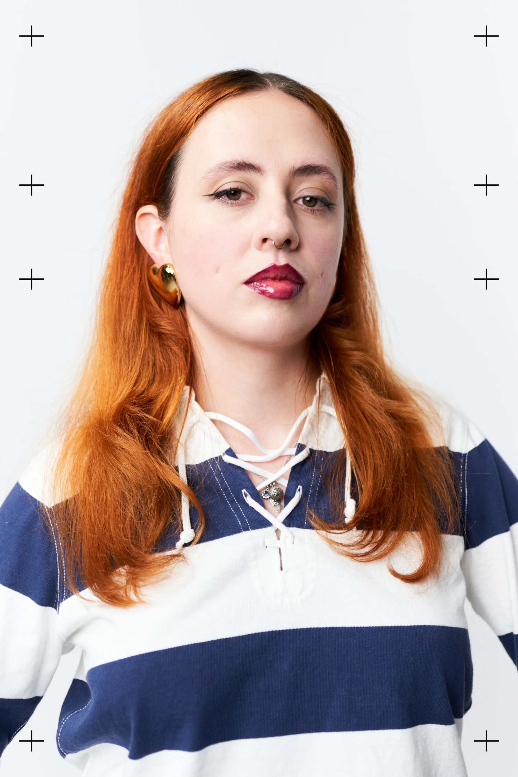 Joven con cabello largo y ondulado de color rojo, vistiendo una camisa a rayas azul marino y blanca, mostrando una expresión de confianza, con pendientes de oro y un collar con colgante.