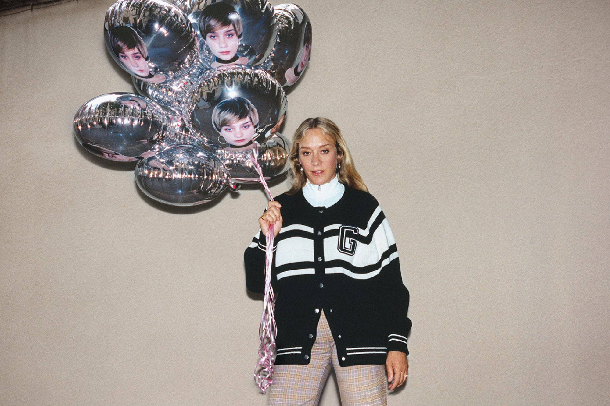 A person holds a cluster of metallic balloons featuring faces, wearing a black and white sweater over a white top and plaid pants.