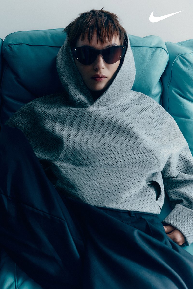 Een persoon met kort haar draagt een zonnebril en een grijze hoodie terwijl hij op een turquoise bank ontspant, met een relaxte uitstraling. Nike-logo zichtbaar.